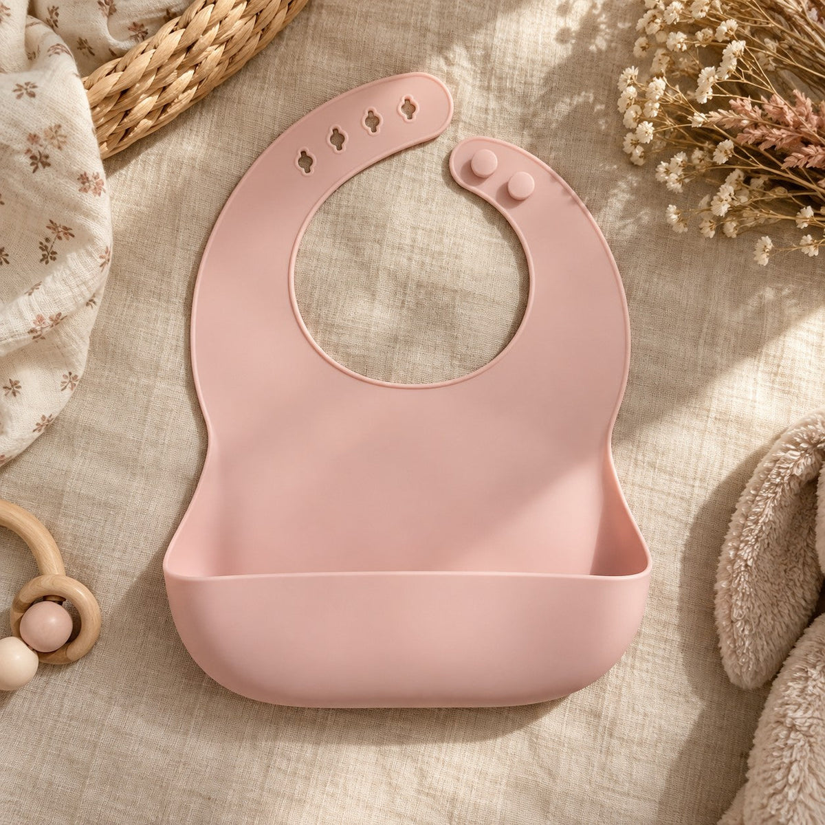 Le bavoir à récupérateur en silicone rose poudré, posé à plat sur un lin beige
