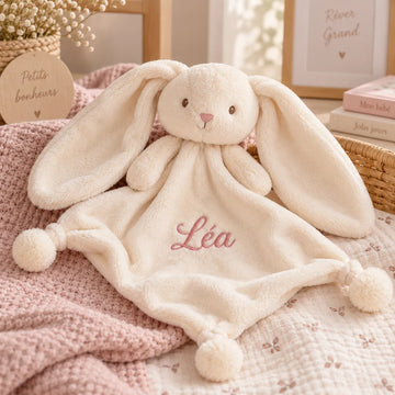 Doudou Lapin Pompon crème brodé au prénom Léa en fil rose, posé sur une couverture